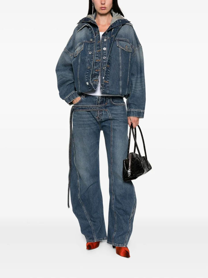 Person wearing a blue denim jacket and jeans holding a black handbag on a white background