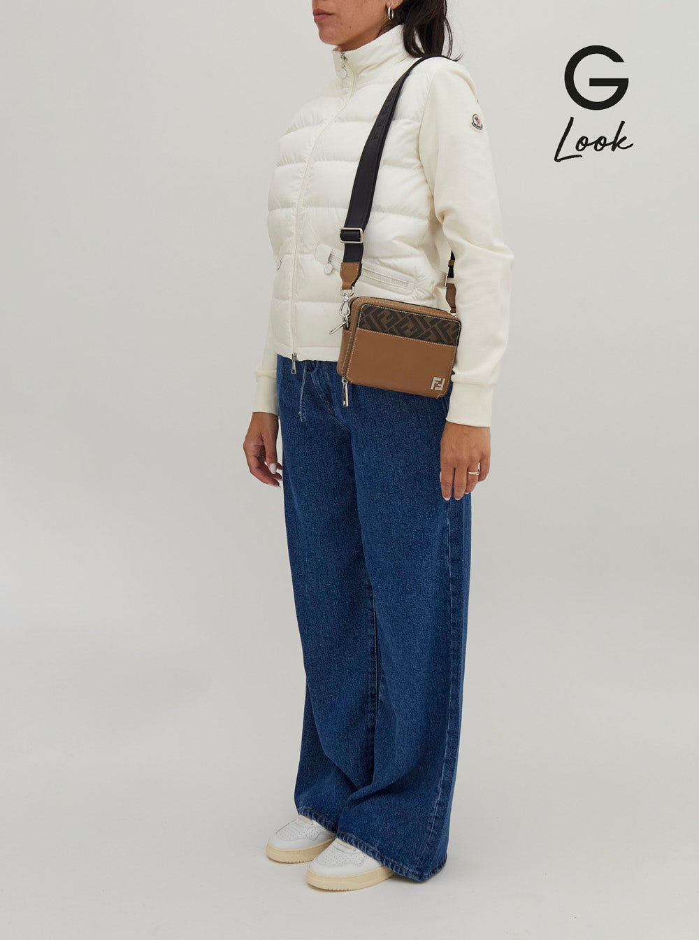 Person wearing a white puffer jacket, blue jeans, and a brown bag with a visible brand logo on a plain background.