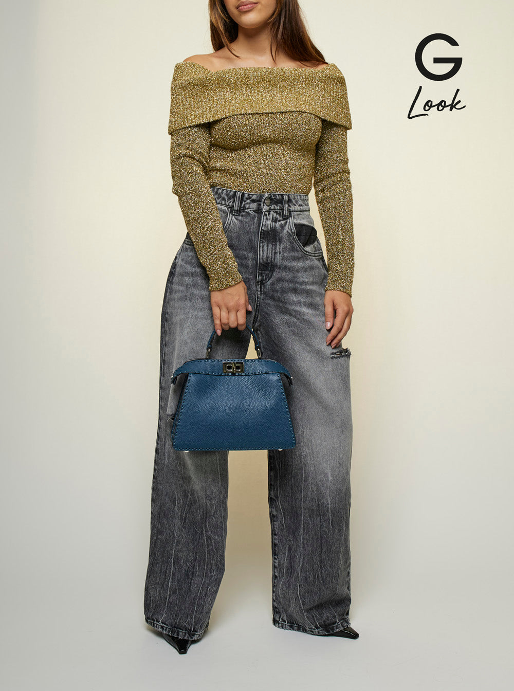 Person wearing a green off-shoulder top and gray jeans, holding a blue handbag on a white background.