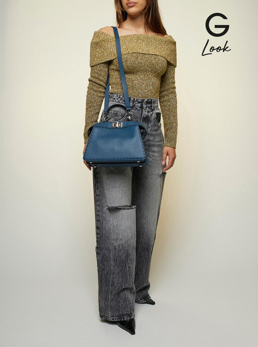 Person wearing a green sweater and gray jeans holding a blue handbag on a white background with 'G Look' branding.