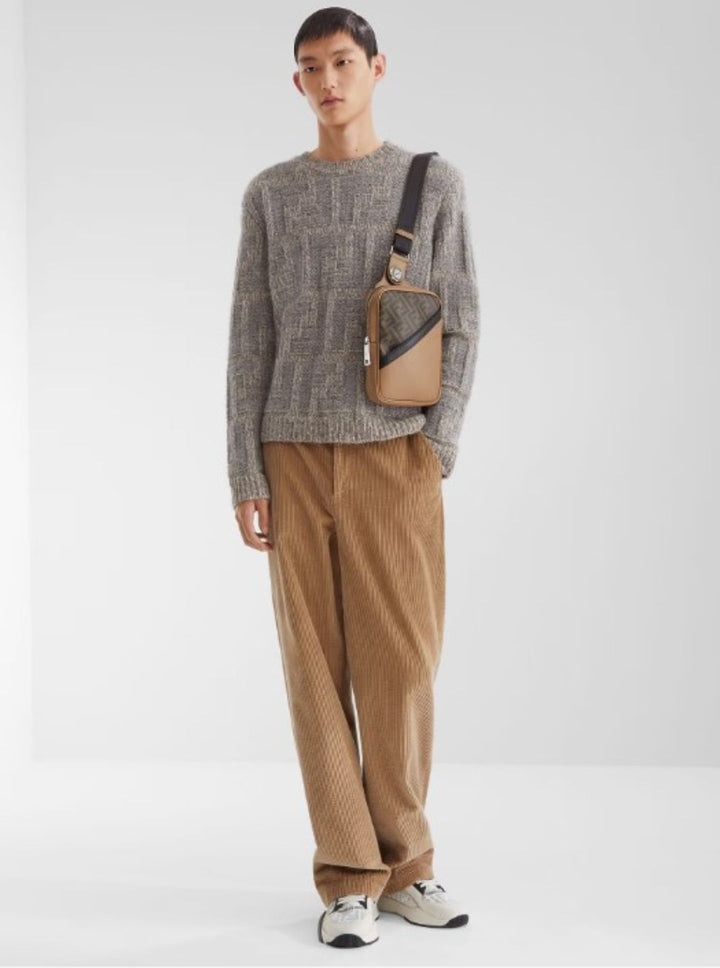 Man wearing a gray sweater and brown pants with a crossbody bag on a white background
