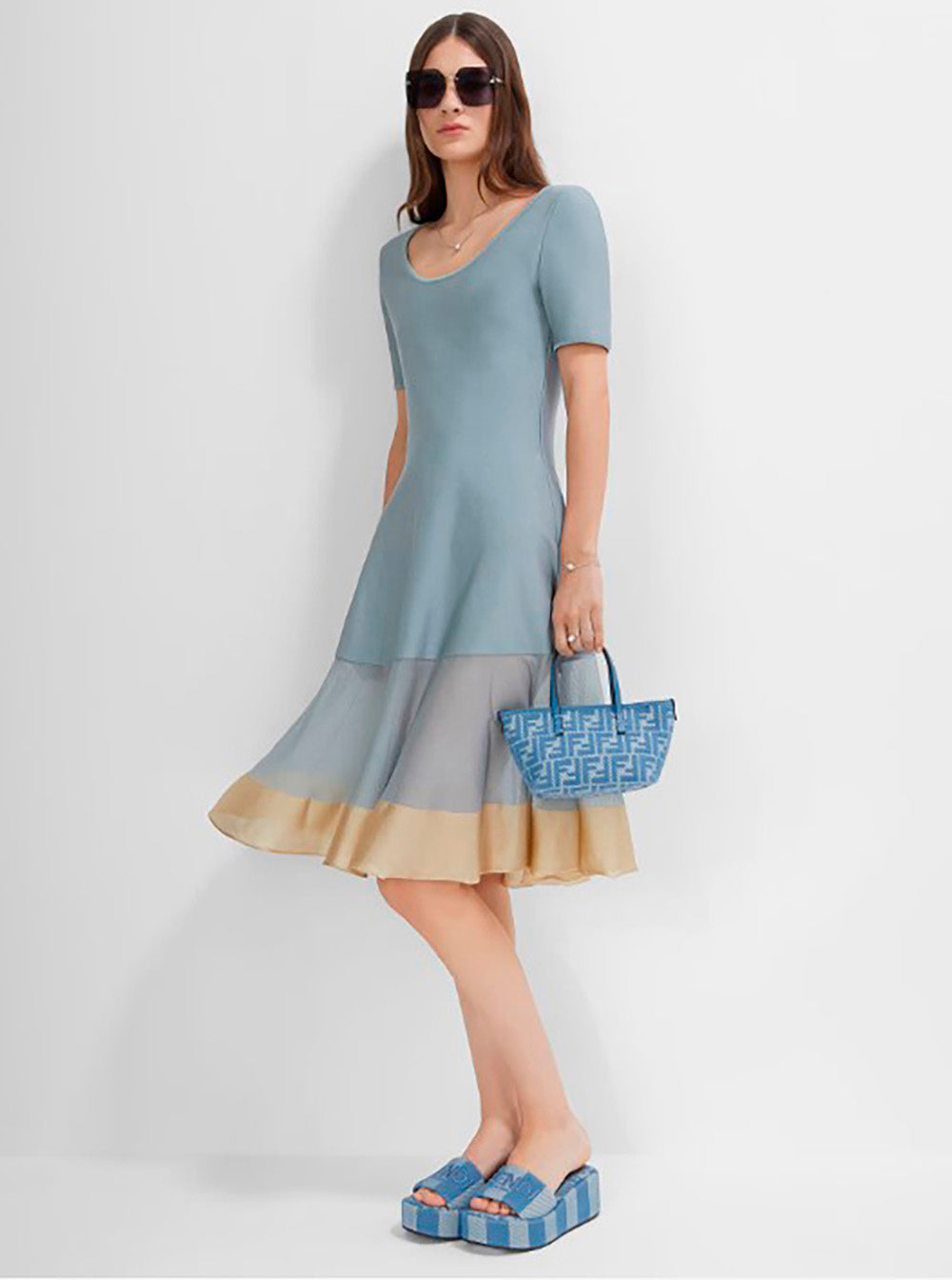 Woman wearing a light blue dress with a sheer overlay, holding a matching handbag against a white background.