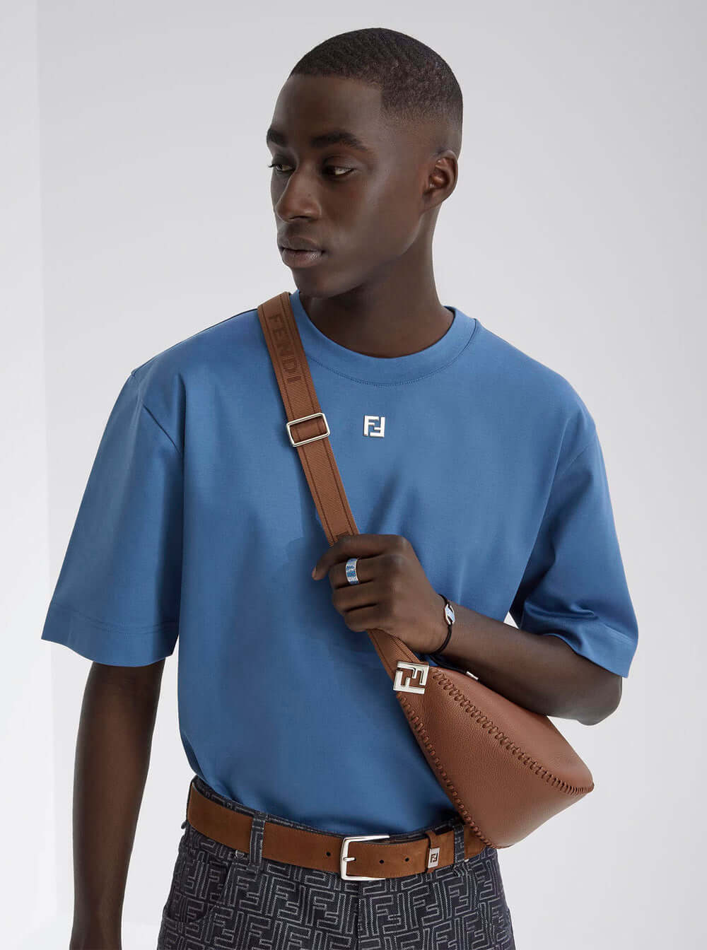 Man wearing a blue t-shirt with a brand logo, holding a brown leather bag against a white background