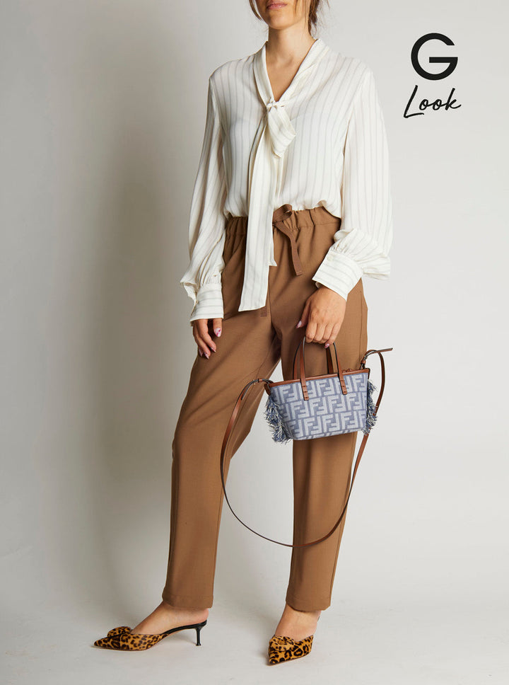 Person wearing a white blouse and brown pants holding a patterned handbag against a plain background.