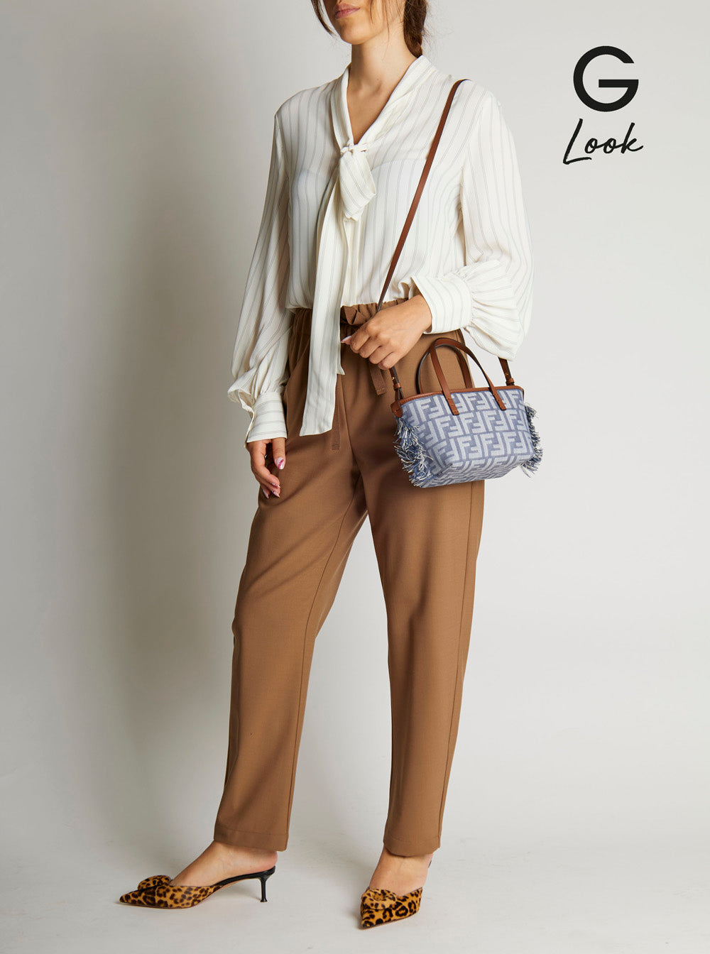 Woman wearing a white blouse and brown pants with a patterned handbag on a plain background