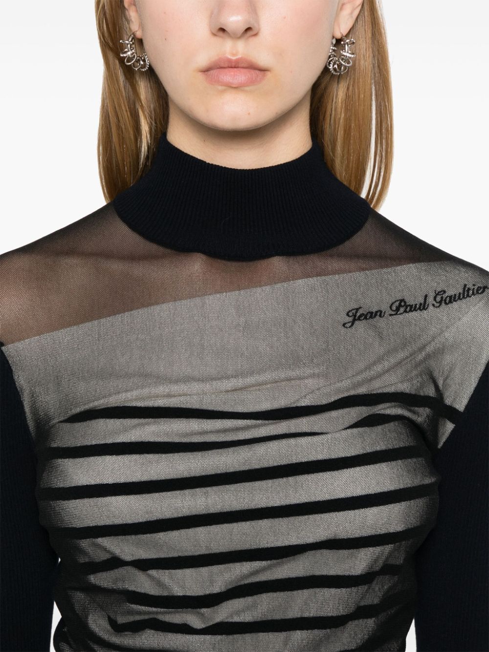 Close-up of a person wearing a black turtleneck and a gray sweater with black stripes, featuring the 'Jean Paul Gaultier' brand.