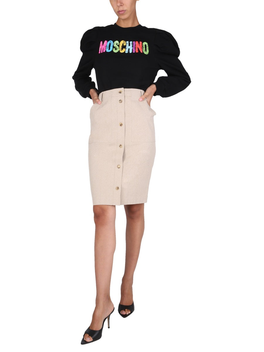 Person wearing a black sweater with colorful branding and a beige button-up skirt on a white background