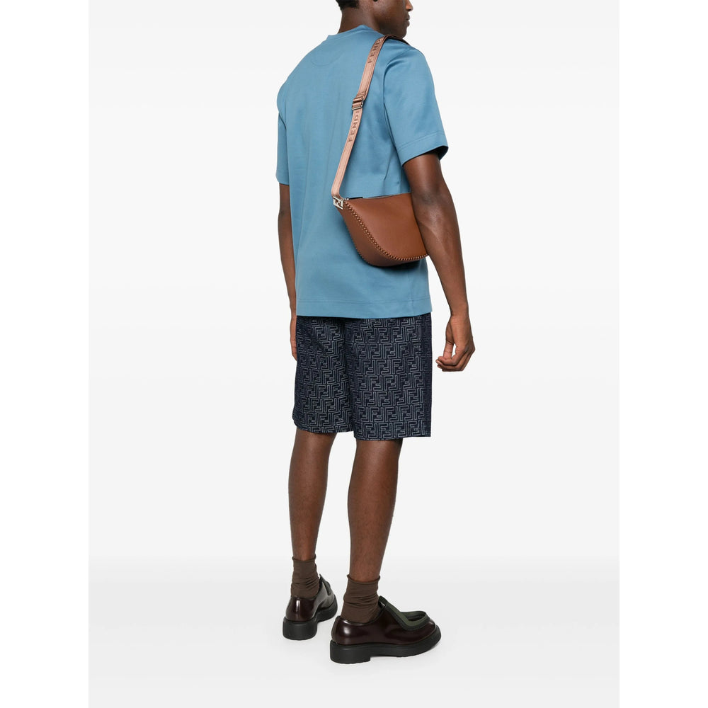 Person wearing a blue shirt, dark shorts, and black shoes with a brown bag over one shoulder on a white background.