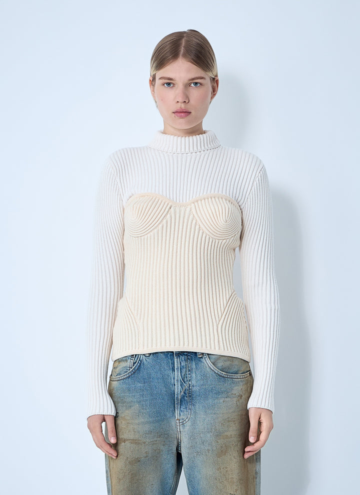 Woman wearing a cream ribbed sweater with a unique design and blue jeans against a light blue background