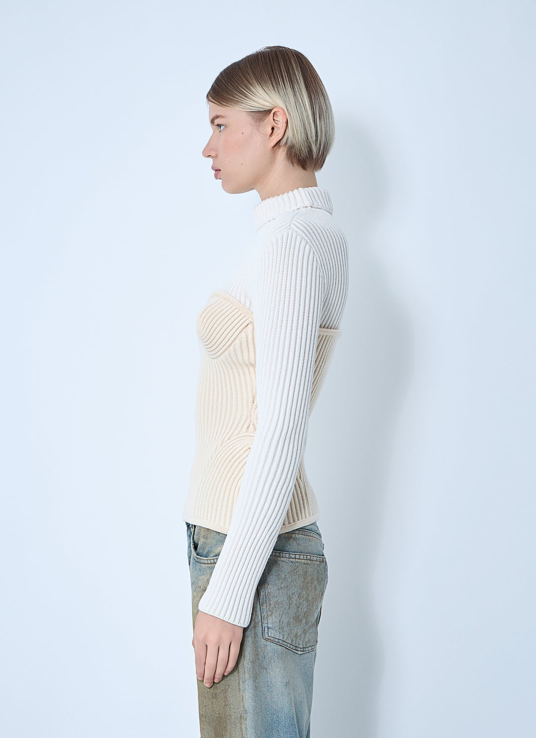 Person wearing a white ribbed sweater over a beige top with light blue jeans against a plain background