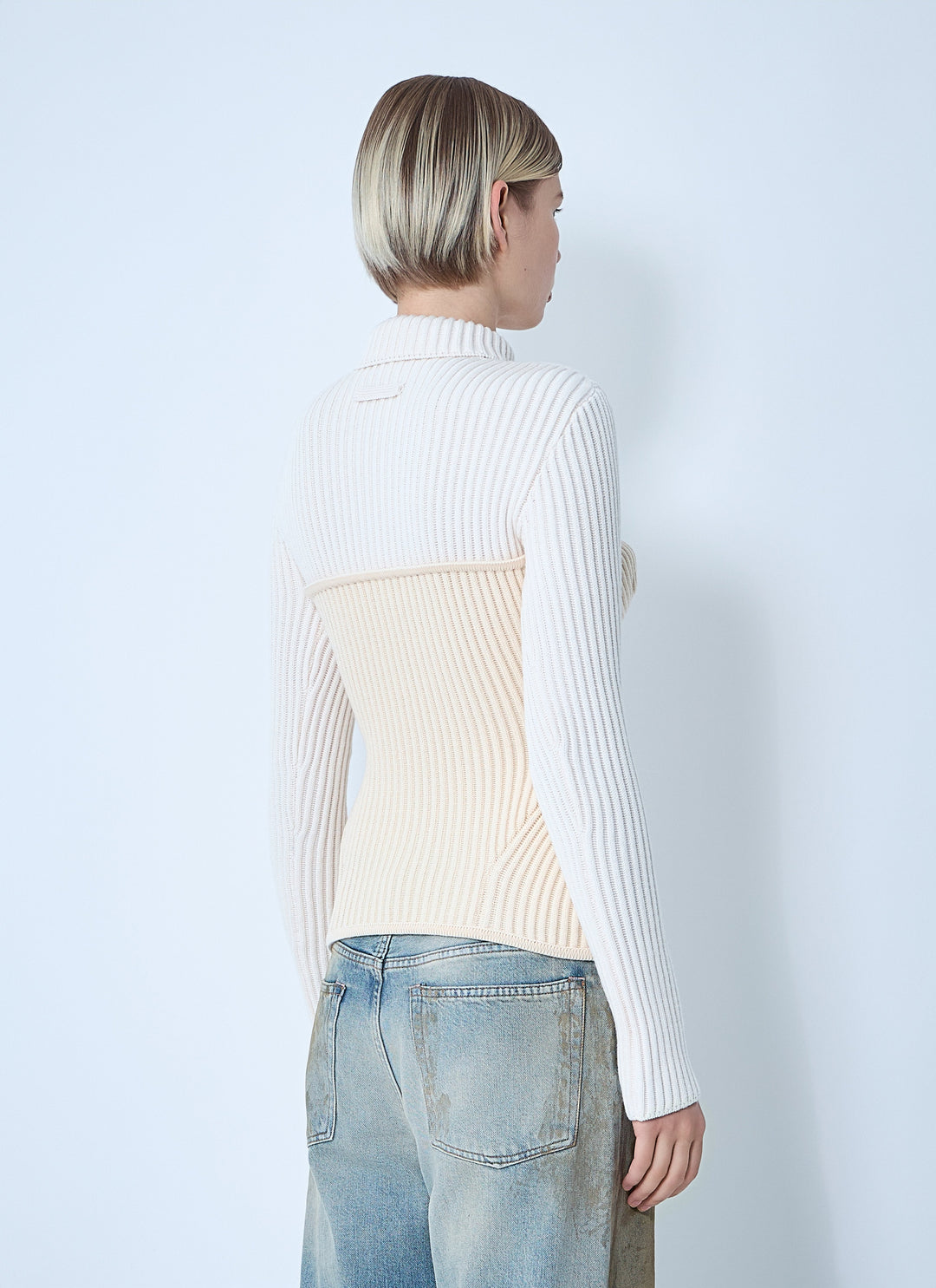 Person wearing a white ribbed sweater over a cream top with light blue jeans on a plain background