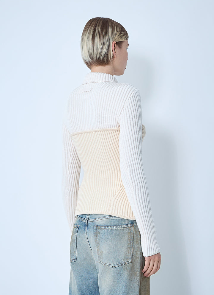 Person wearing a white ribbed sweater over a cream top with light blue jeans on a plain background