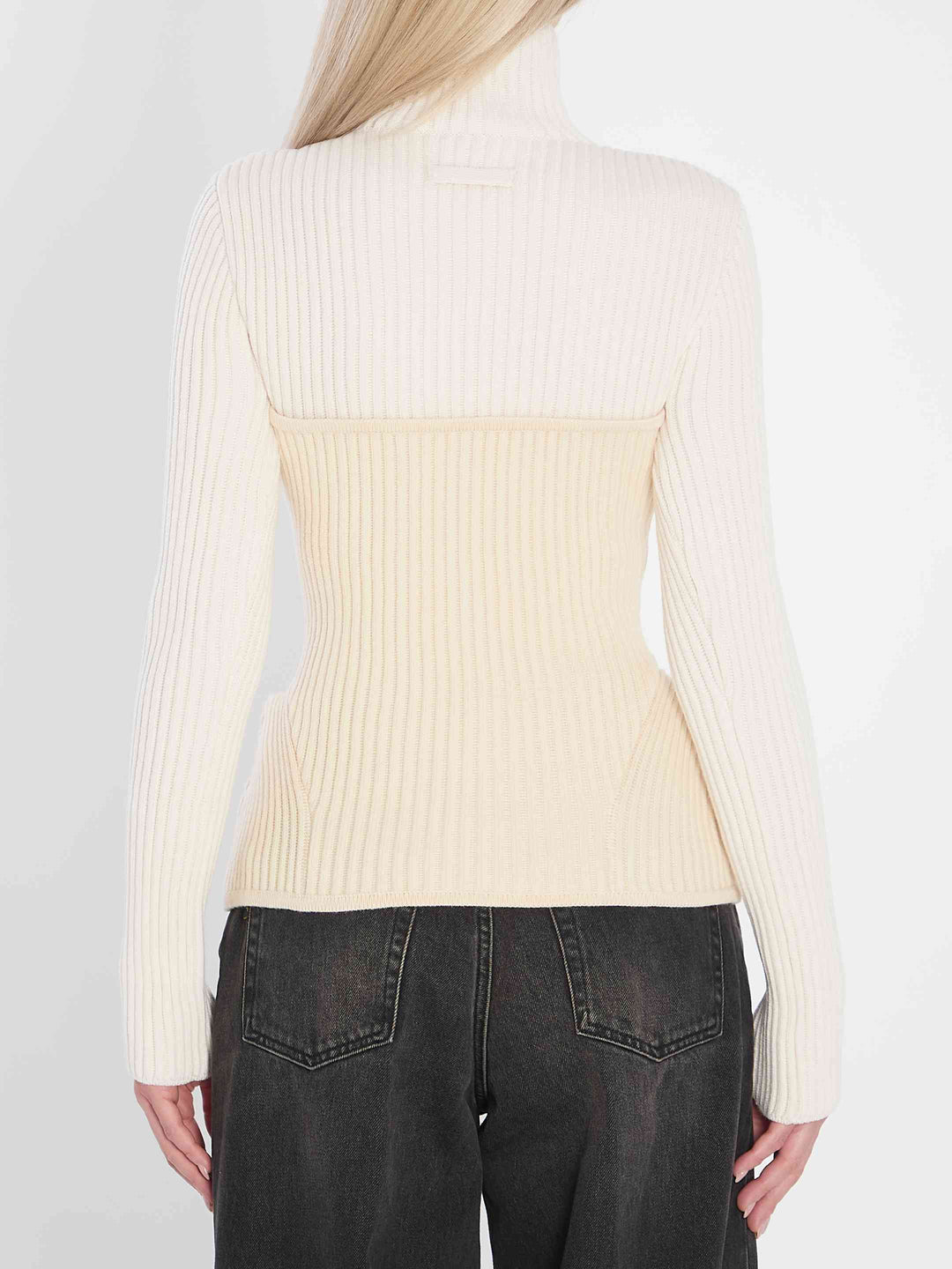 Person wearing a cream ribbed sweater with a color-block design, paired with dark jeans on a plain background.