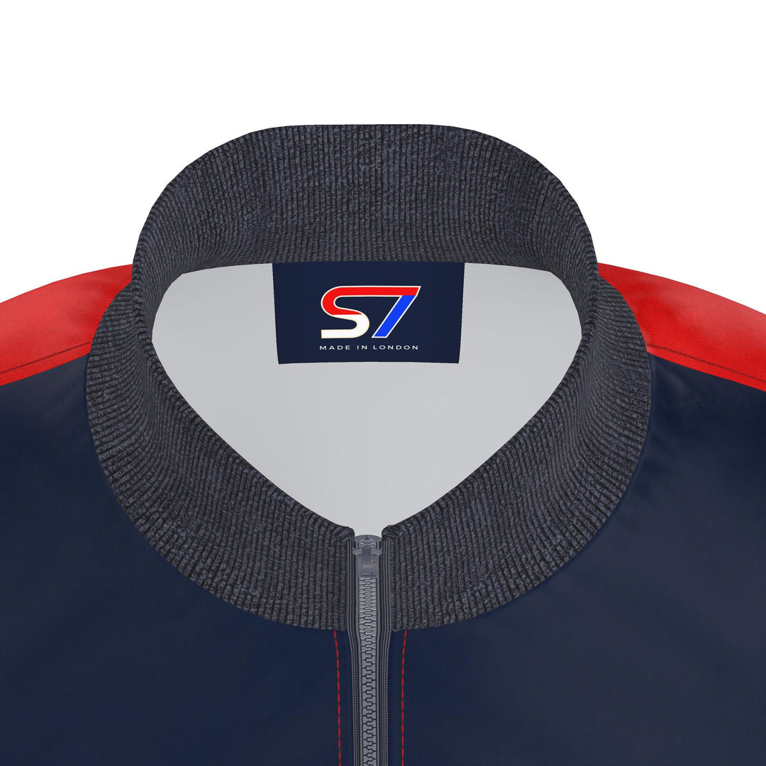 Close-up of a jacket collar and upper chest showing a dark gray ribbed knit collar, a navy body, red shoulder panels, and a central logo patch reading S7 MADE IN LONDON.