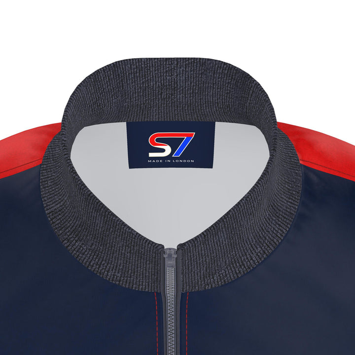 Close-up of a jacket collar and upper chest showing a dark gray ribbed knit collar, a navy body, red shoulder panels, and a central logo patch reading S7 MADE IN LONDON.