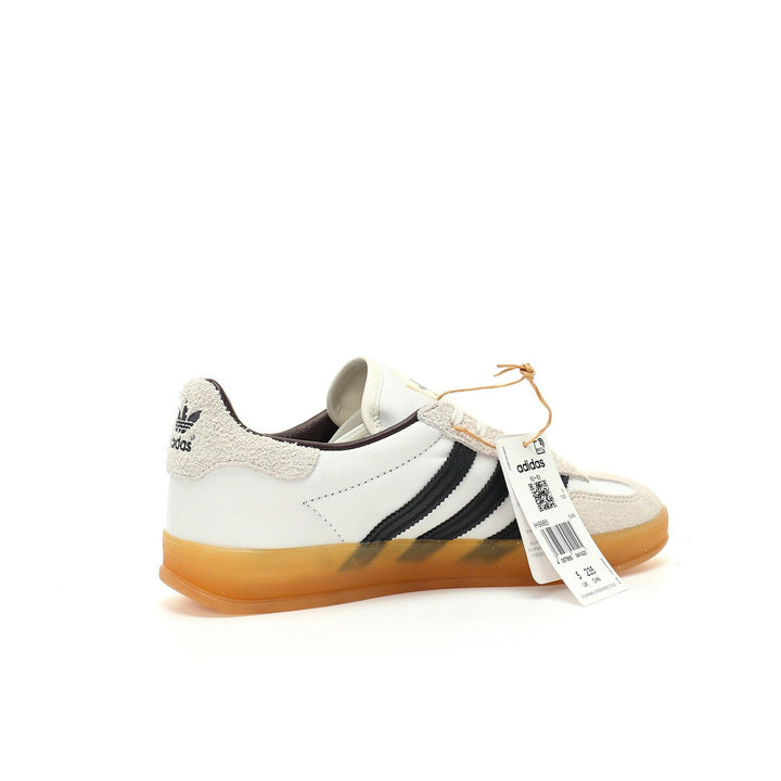 A white Adidas Gazelle sneaker with black three-stripes, a beige suede toe cap, and a translucent gum sole, shown with its retail tag attached on a white background.