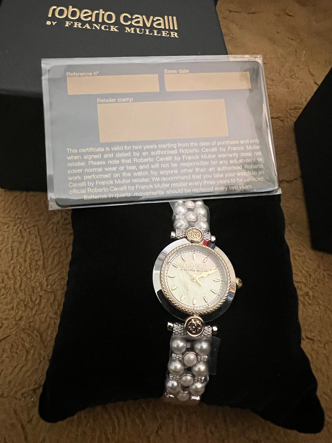 A Roberto Cavalli by Franck Muller watch displayed on a black cushion, with its certificate of authenticity in a clear plastic sleeve and part of the black branded box visible behind it.