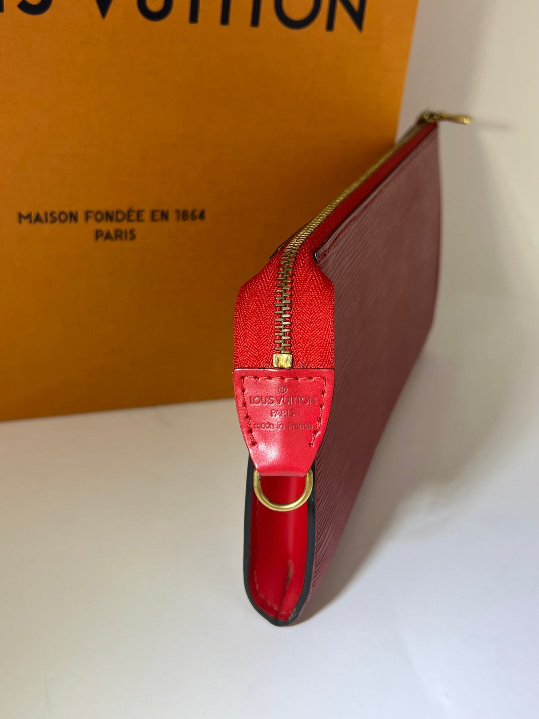 Red wallet with gold zipper and hardware on a white surface, with a Louis Vuitton box in the background.