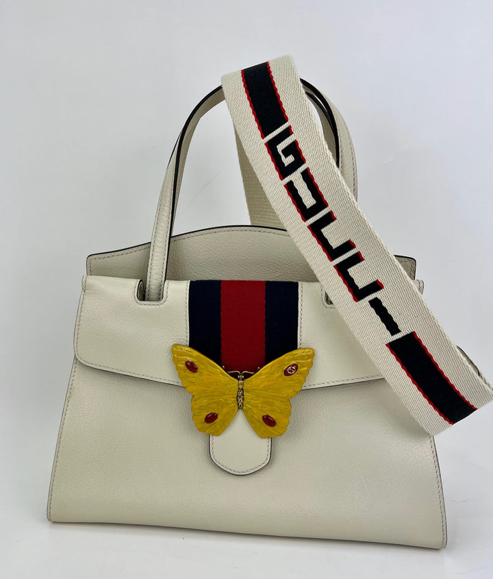 White handbag with a butterfly charm and a branded strap on a white background