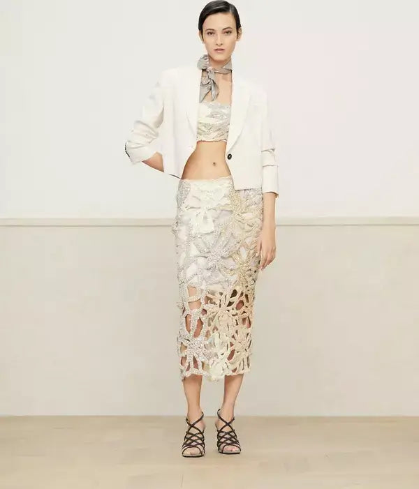 Woman wearing a white blazer, lace top, and lace skirt against a plain background