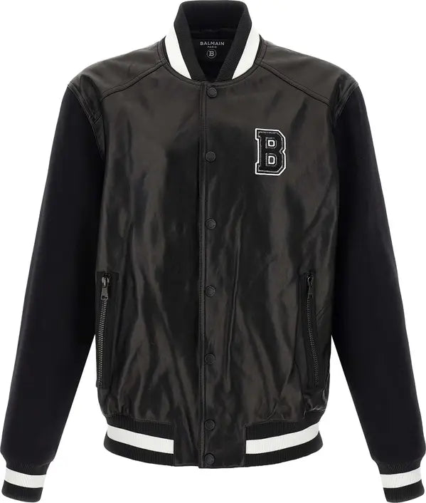 A Balmain black satin bomber jacket with black ribbed sleeves, collar, and cuffs, accented by white stripes on the collar, cuffs, and hem. It features a white embroidered &