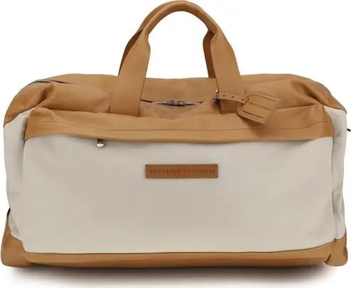 A tan and off-white duffel bag with dual top handles, a zippered top opening, and a small brown leather brand patch on the front that reads 'THE TRAVEL SHOP'.