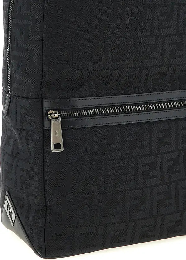 Close-up of a black Fendi backpack featuring the signature FF monogram pattern, a horizontal zippered pocket with a silver zipper pull, and a small Fendi logo tab near the bottom left corner.