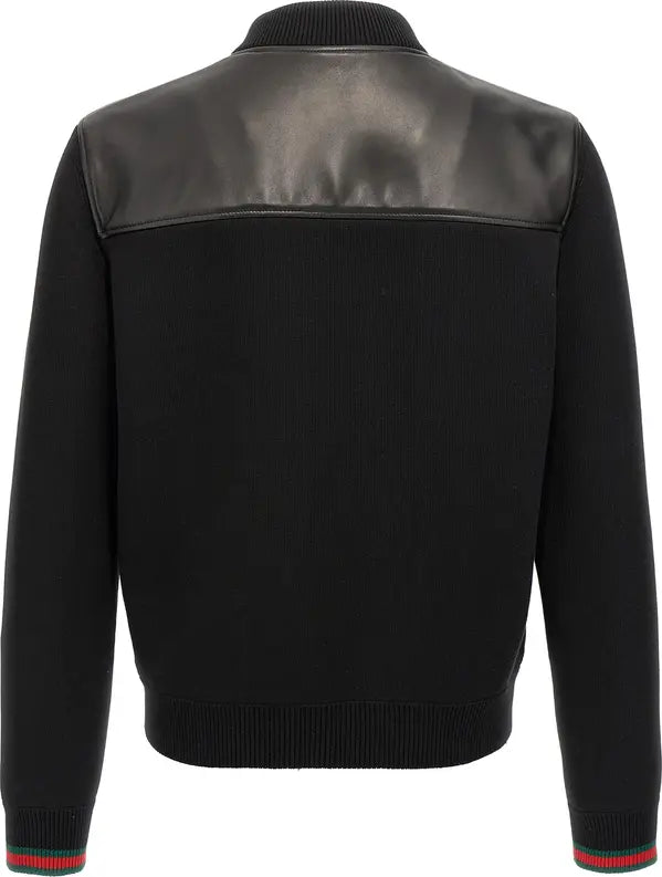 Black knit bomber jacket with black leather shoulder panels, ribbed collar, cuffs, and hem, featuring red and green striped knit trim at the cuffs.