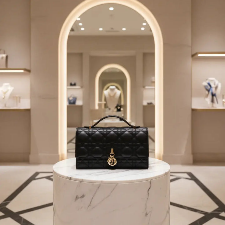 A black quilted leather handbag with a top handle and a gold-tone Christian Dior CD charm on the front flap, set against a plain white background.