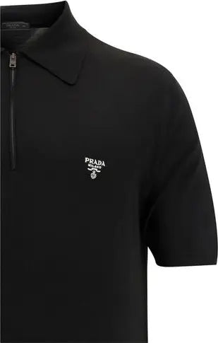 Close-up of a black short-sleeve polo shirt with a half-zip collar and a small white embroidered Prada logo on the left chest, shown against a plain white background.