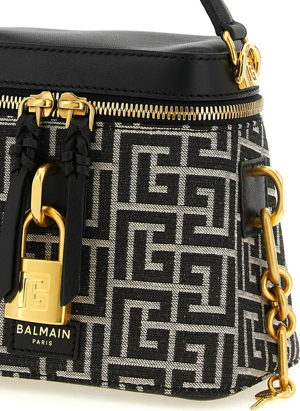 Close-up of a Balmain handbag featuring a black and white Greek key monogram pattern, black leather trim, gold-tone hardware, and a hanging gold padlock charm with the Balmain logo.