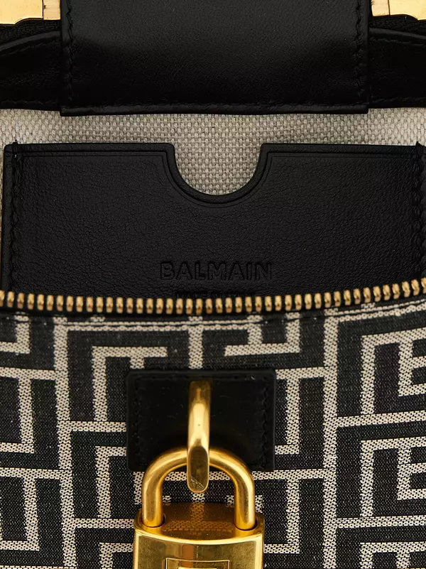 Close-up of a Balmain handbag showing the black leather logo patch, gold zipper, and black-and-white geometric monogram canvas with a gold padlock detail.
