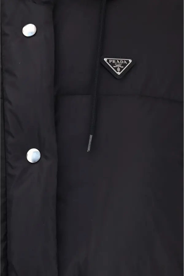 Close-up of a black puffer jacket showing the Prada triangular logo patch on the chest, a drawstring hood, and silver snap buttons on the front.