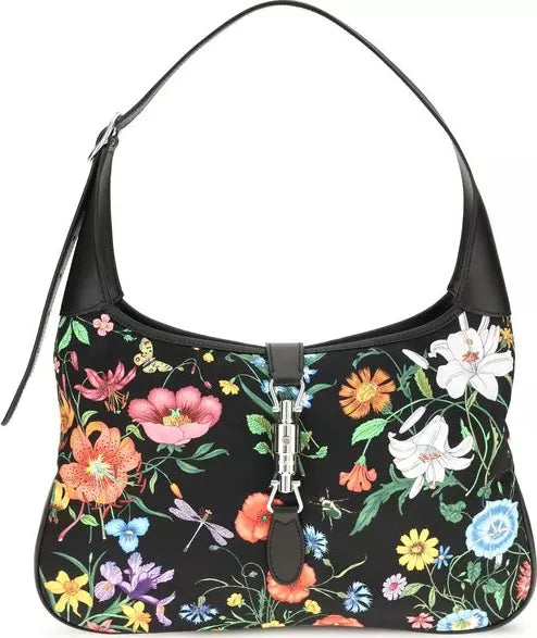 A black shoulder bag with a vibrant multicolor floral print, featuring a central silver-tone turn-lock clasp and a black leather shoulder strap on a white background.