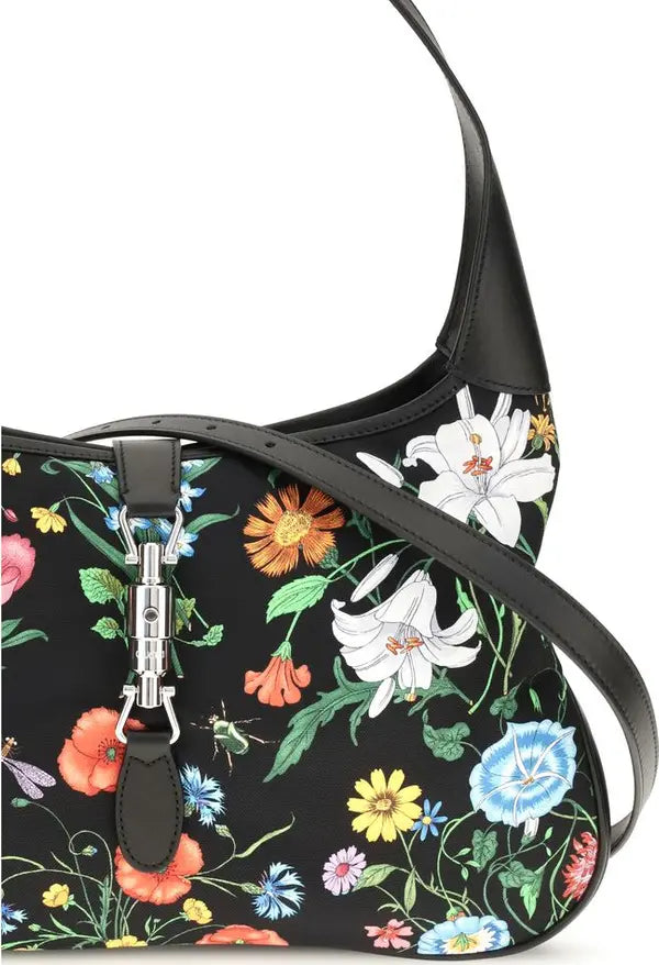 A black shoulder bag with a vibrant multicolor floral print, featuring white lilies, orange poppies, and blue flowers, with black leather trim and a silver-tone clasp.