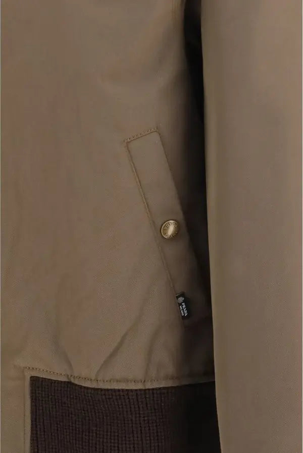 Close-up of a beige jacket with a side flap pocket and a gold-tone snap button, shown on a mannequin or hanger against a matching neutral background.