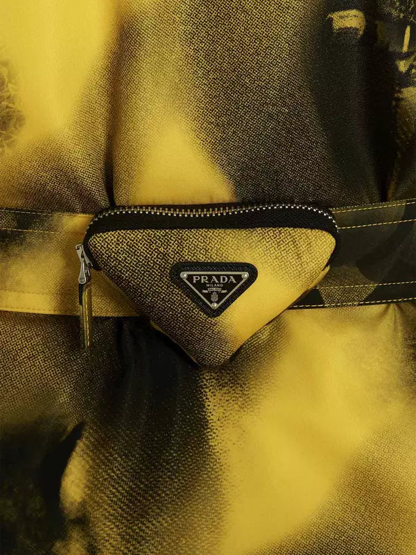 A close-up of a Prada belt bag in a textured canvas material, featuring the signature triangular Prada logo plaque on the front. The bag is styled on a matching Prada belt with a metallic gold finish, set against a background of draped gold fabric.