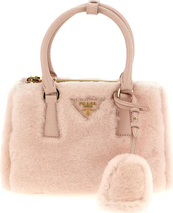 A light pink Prada handbag made of soft faux fur, featuring a structured top handle, a matching fur charm, and the signature gold-tone triangular Prada logo plaque on the front.