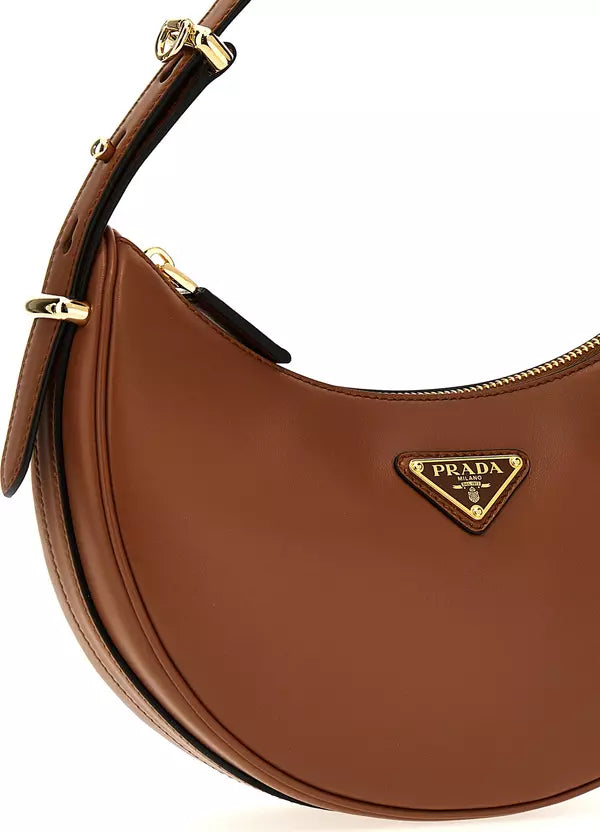A close-up of a brown leather Prada crescent shoulder bag with gold-tone hardware and the signature triangular Prada logo plaque on a white background.