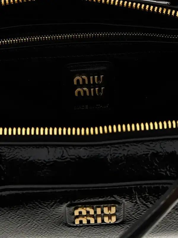Close-up of a black textured leather handbag interior with gold-tone zippers and a black Miu Miu logo plaque on the inside, plus a matching Miu Miu logo plaque on the exterior.