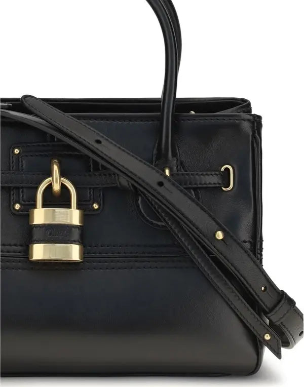A close-up of a black leather handbag featuring a prominent gold padlock closure on the front and a matching black shoulder strap.