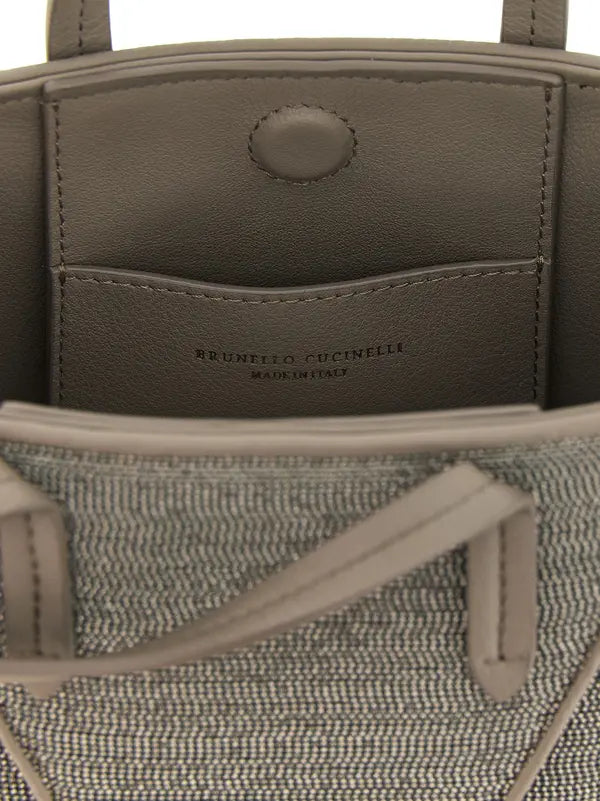 Close-up of the interior of a Brunello Cucinelli handbag, showing a taupe leather pocket with the brand name debossed, a textured gray and white woven fabric body, and taupe leather handles.