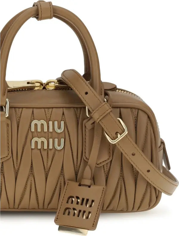 A Miu Miu taupe leather handbag with a top handle, gold-tone hardware, and a matching leather tag featuring the Miu Miu logo.