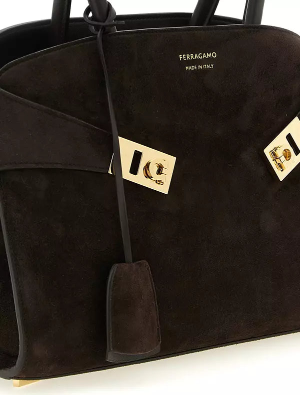 A close-up of a black suede Ferragamo handbag with gold-tone hardware and a hanging clochette, on a white background.