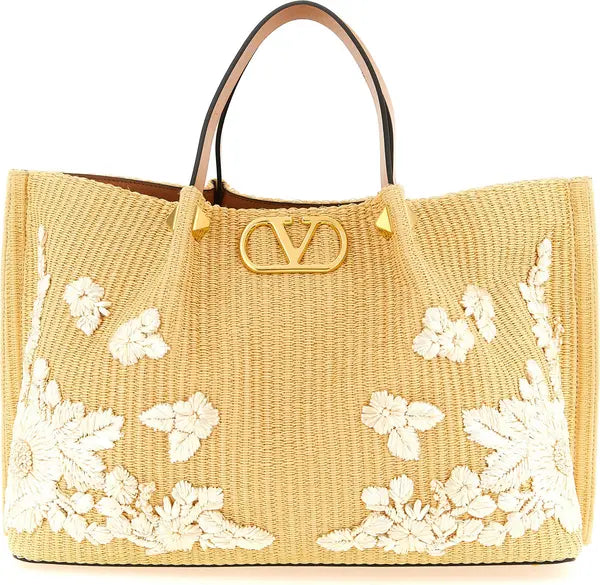 A beige woven straw tote bag with white floral embroidery on the front, featuring a gold-tone metal V logo clasp and dark brown leather handles, photographed on a solid white background.