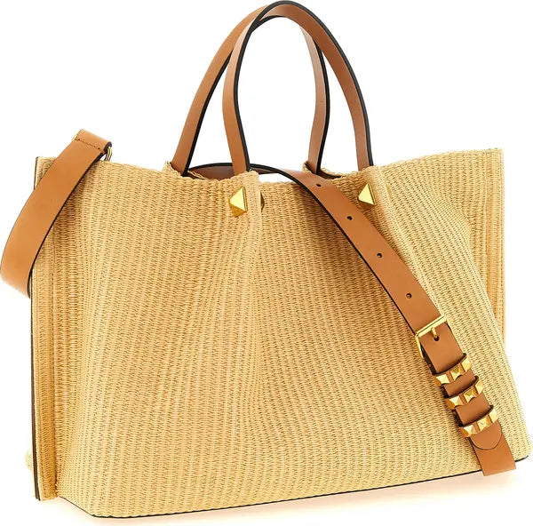A woven straw tote bag in a light tan color with brown leather handles, a matching shoulder strap, and gold-tone metal studs and buckles, shown on a plain white background.