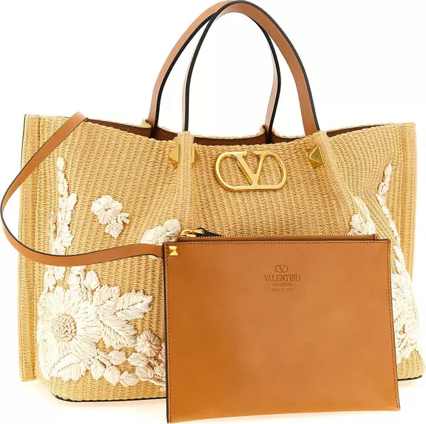 A beige woven tote bag with white floral embroidery, tan leather handles and shoulder strap, a gold-tone V logo clasp, and a matching tan leather zip pouch in front. The Valentino logo is embossed on the pouch.