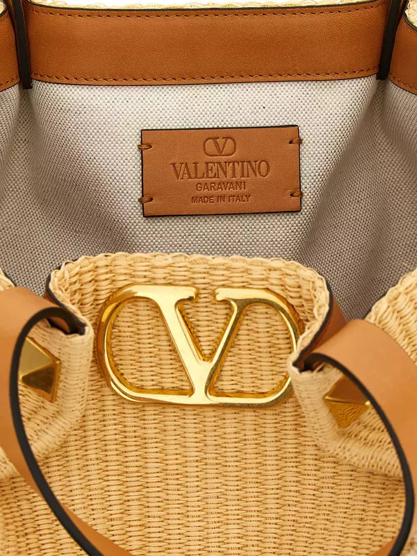 Close-up of a Valentino Garavani woven straw bucket bag interior, showing a tan leather interior label reading Valentino Garavani Made in Italy, tan leather trim, and a large gold-tone metal VC logo clasp on the woven front.