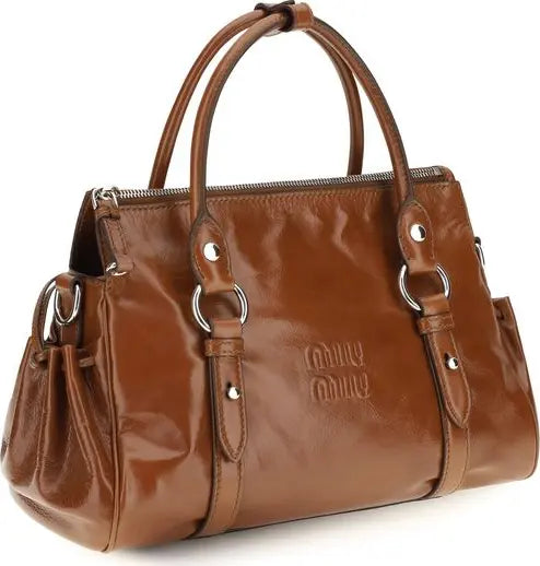 A brown patent leather handbag with dual top handles, silver-tone hardware, and an embossed logo on the front, shown against a plain white background.