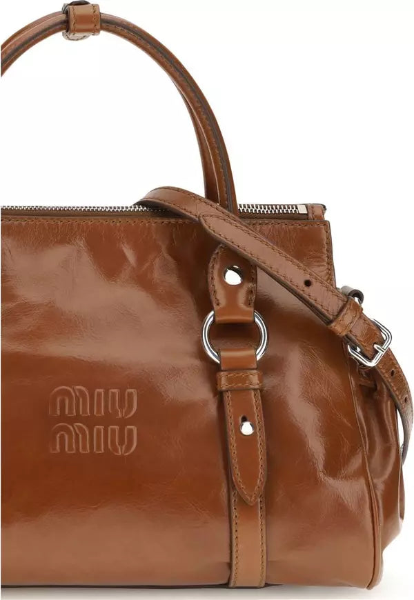 A close-up of a brown leather Miu Miu handbag with a glossy finish, featuring a top handle, a shoulder strap with silver-tone hardware, and the debossed Miu Miu logo on the front.